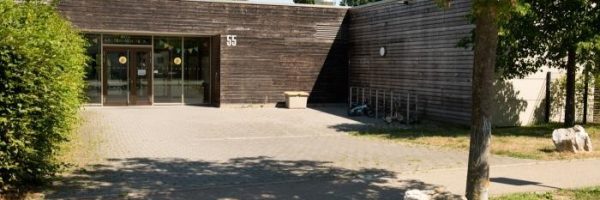 Eine moderne, einstöckige Kita in der Lautenbacher Straße mit Holzverkleidung und großen Glastüren, umgeben von Bäumen und einem gepflasterten Innenhof. Die Nummer 55 steht an der Fassade, in der Nähe des Eingangs befinden sich Fahrradständer und Bänke.