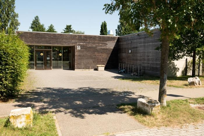 Eine moderne, einstöckige Kita in der Lautenbacher Straße mit Holzverkleidung und großen Glastüren, umgeben von Bäumen und einem gepflasterten Innenhof. Die Nummer 55 steht an der Fassade, in der Nähe des Eingangs befinden sich Fahrradständer und Bänke.