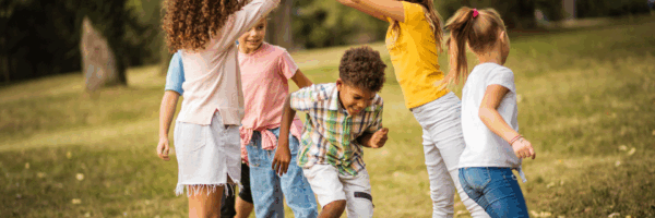 Sechs Kinder spielen zusammen in einem Park mit Gras, bilden mit ihren Armen einen Bogen, während sich ein Kind darunter duckt, alle lächeln und genießen die Natur mit den Bäumen im Hintergrund.