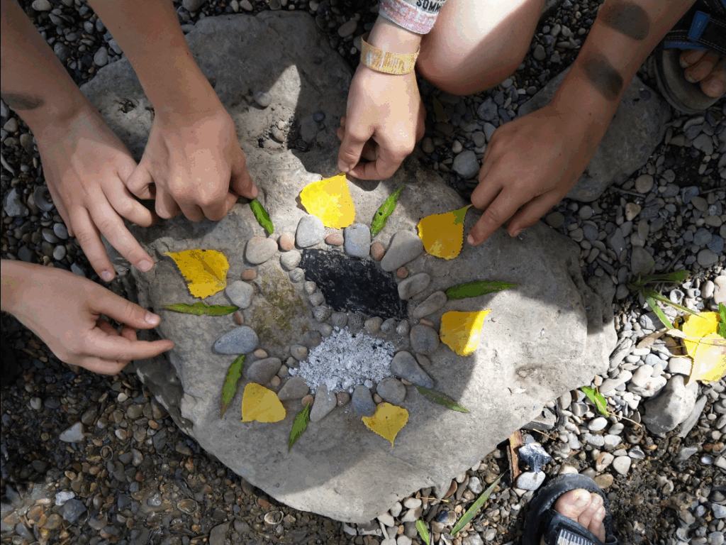 Vier Kinderhände ordnen gelbe Blütenblätter, grüne Blätter und kleine Steine in einem kreisförmigen Muster auf einem großen Felsen an und schaffen so Naturkunst im Freien auf einem kiesigen Boden.