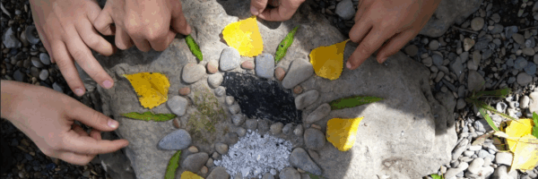 Vier Kinderhände ordnen gelbe Blütenblätter, grüne Blätter und kleine Steine in einem kreisförmigen Muster auf einem großen Felsen an und schaffen so Naturkunst im Freien auf einem kiesigen Boden.