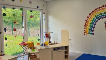 Ein helles Klassenzimmer im PARI Kinderhaus mit einem Spielküchen-Set, einem blauen Teppich und bunten Punkten, die einen Regenbogen bilden. Große Fenster lassen natürliches Licht herein, und draußen sind Bäume zu sehen, die eine fröhliche Kindertageseinrichtung schaffen.