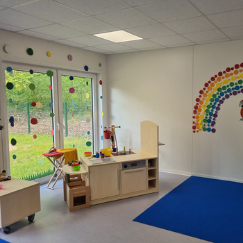 Ein helles Klassenzimmer im PARI Kinderhaus mit einem Spielküchen-Set, einem blauen Teppich und bunten Punkten, die einen Regenbogen bilden. Große Fenster lassen natürliches Licht herein, und draußen sind Bäume zu sehen, die eine fröhliche Kindertageseinrichtung schaffen.