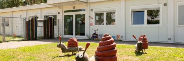 Drei große, bunte Schneckenskulpturen mit roten Häusern zieren das Gras vor einer Kindertageseinrichtung in der Pichterichstraße, einem weißen einstöckigen Gebäude mit großen Fenstern. Neben dem Eingang steht ein Fahrrad, eingerahmt von Bäumen und blauem Himmel.